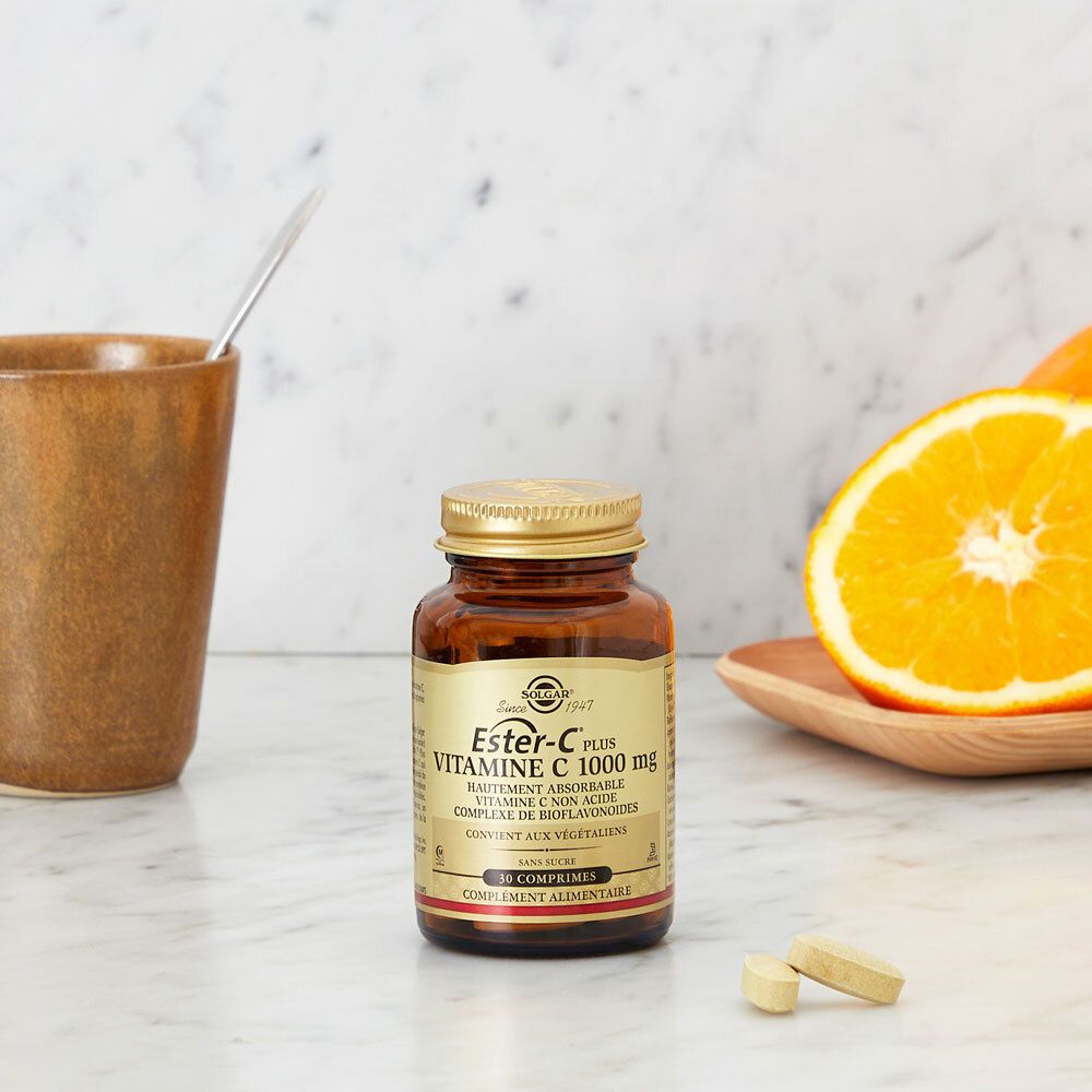 Flacone con compresse, tazza e frutta. Nome prodotto: Ester-C Plus 1000 mg. Su un tavolo.