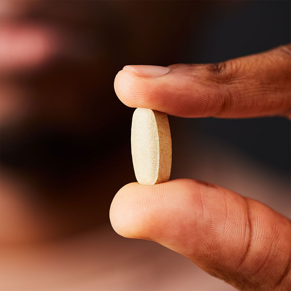 Persona che tiene una compressa tra pollice e indice. Colore beige chiaro. Bordi arrotondati.