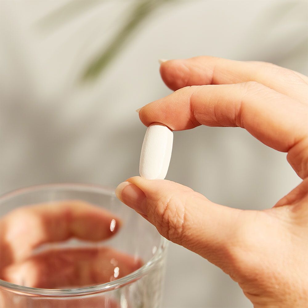 Mano che tiene una compressa bianca ovale sopra un bicchiere d'acqua.