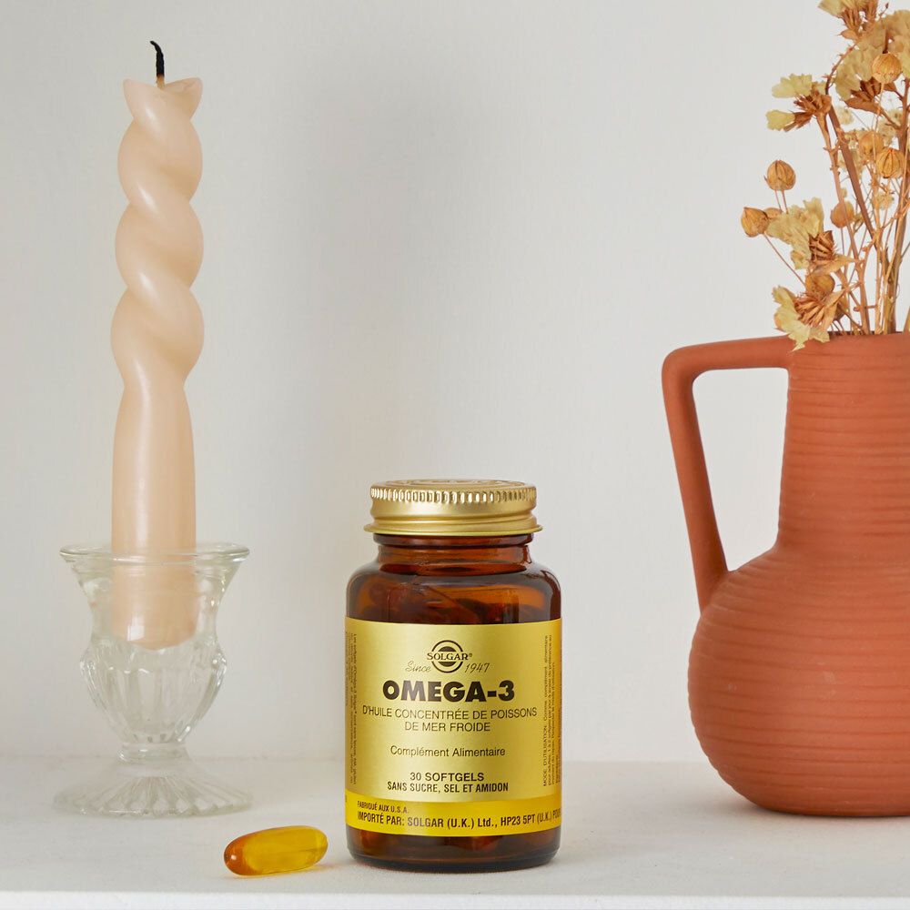 Flacone marrone con tappo dorato. Vicino a una candela e un vaso. Una capsula accanto.