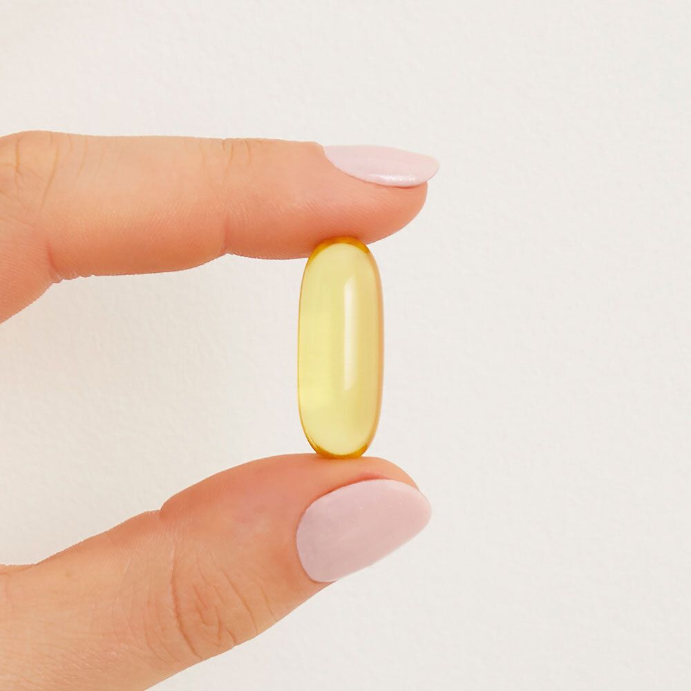 Una singola softgel gialla, tenuta tra le dita. Forma ovale trasparente.