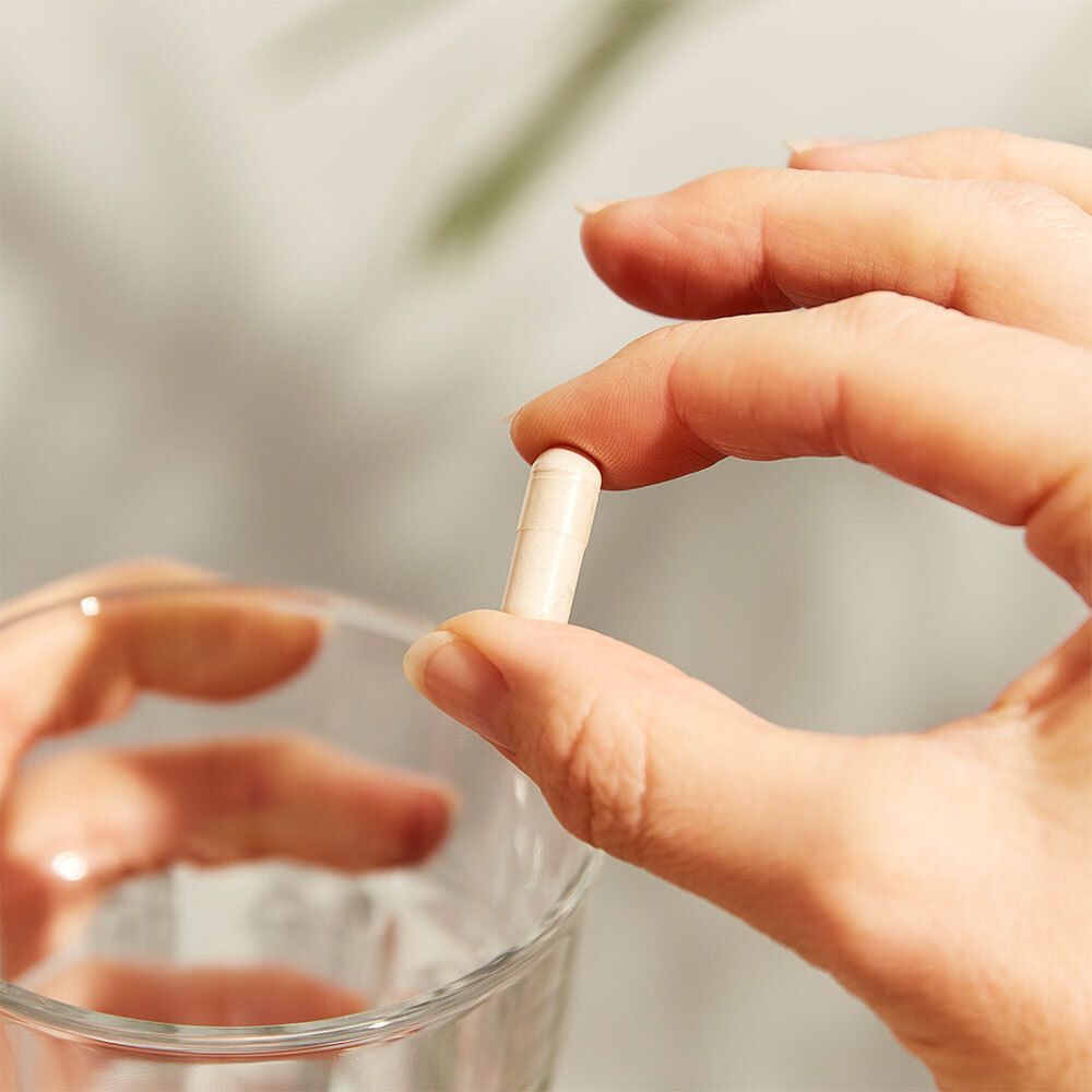 Mano che tiene una capsula sopra un bicchiere d'acqua. Pelle chiara, sfondo sfocato.