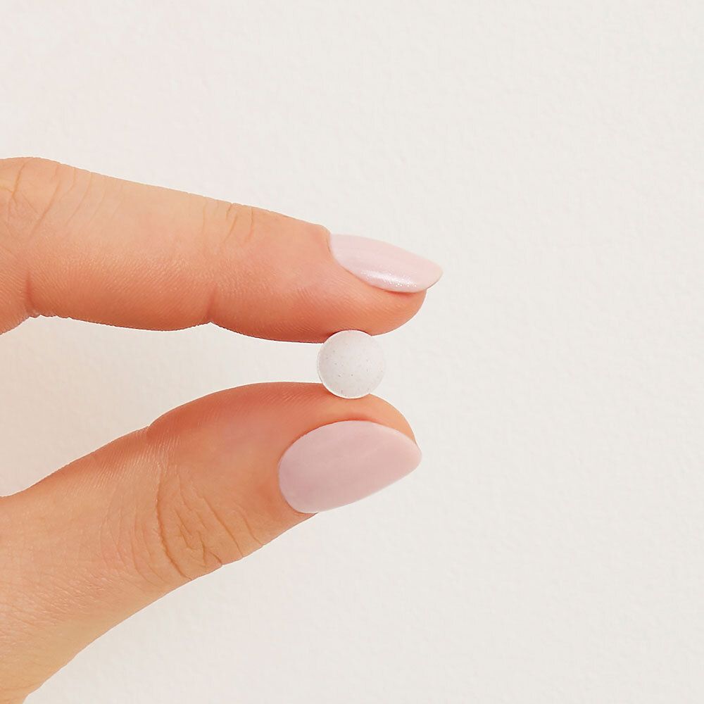 Una singola compressa rotonda e bianca, tenuta tra le dita con smalto rosa.