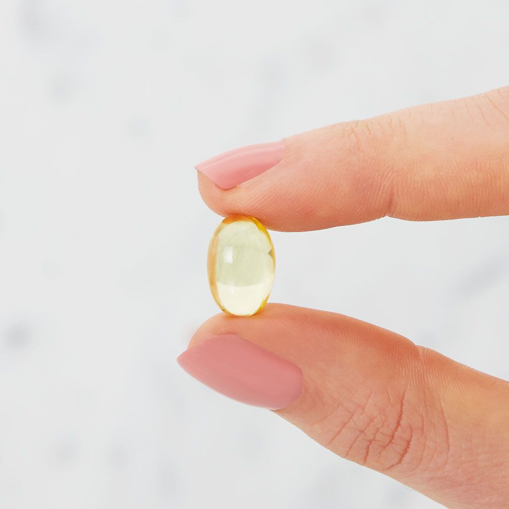 Primo piano di una capsula di gelatina morbida ovale gialla, tenuta tra le dita. Sfondo chiaro e sfocato.