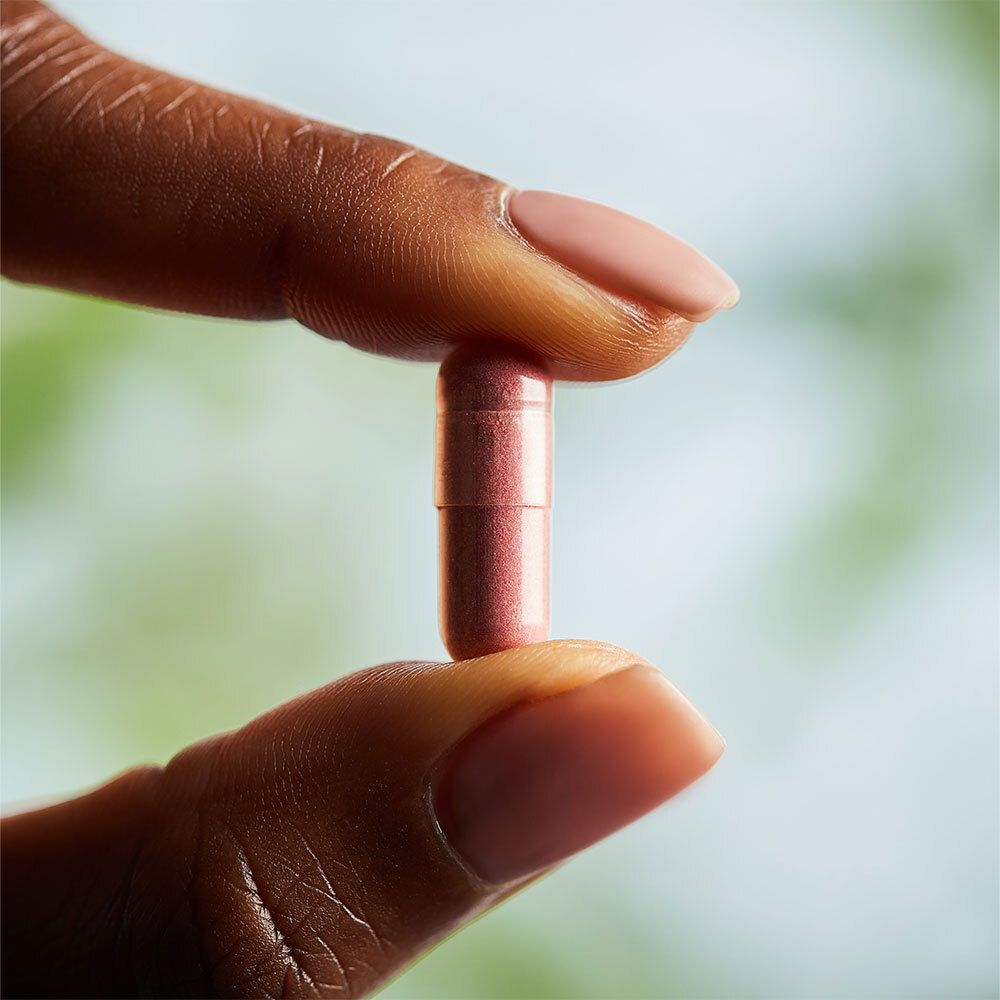 Una capsula rosa tenuta tra due dita. Sfondo: toni verdi e blu sfocati.