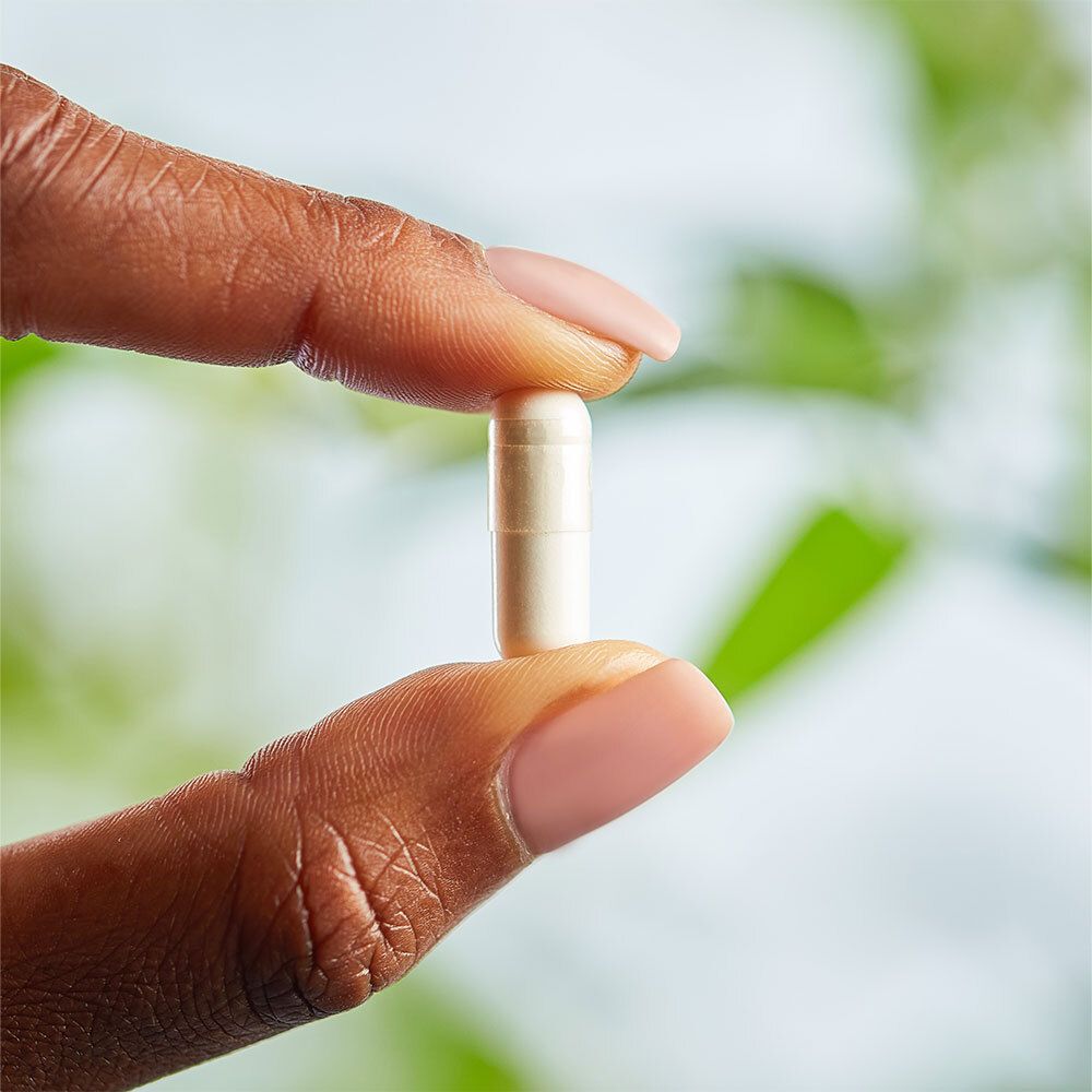 Una mano tiene una capsula. La capsula è bianca e traslucida. Sfondo: fogliame verde.