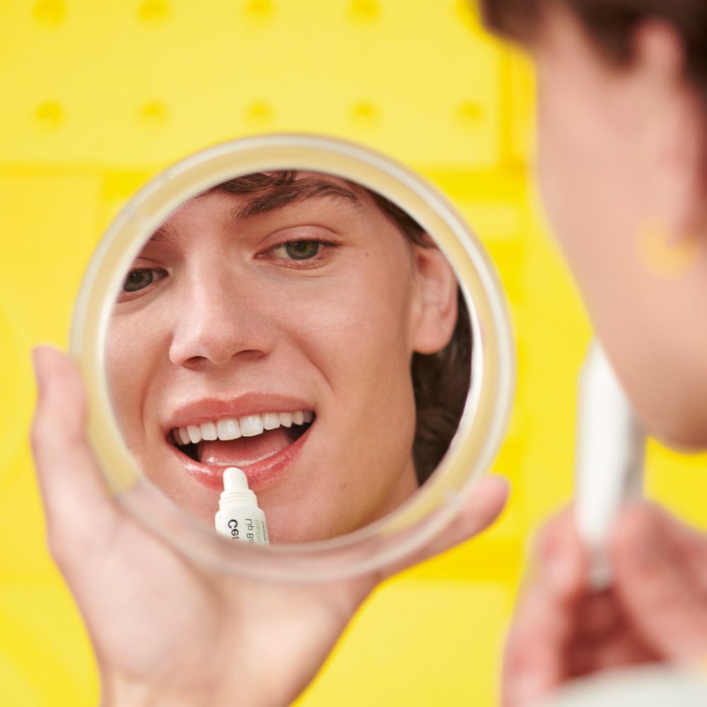Persona si guarda allo specchio e applica balsamo labbra. Sfondo giallo.