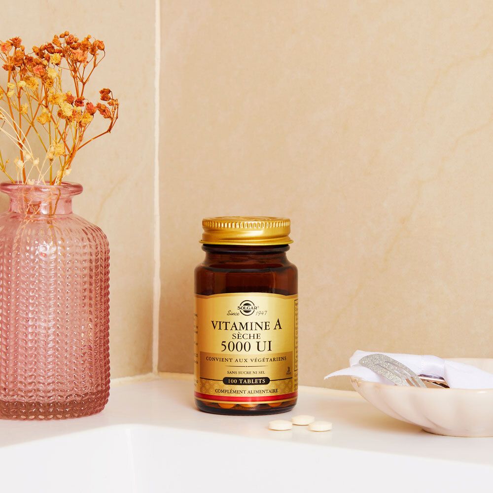 Flacone marrone con tappo dorato su una superficie bianca. Accanto, un vaso con fiori.