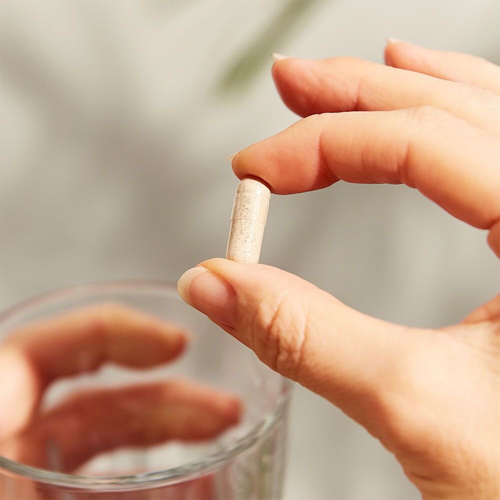 Una mano tiene una capsula sopra un bicchiere d'acqua. La capsula è beige.