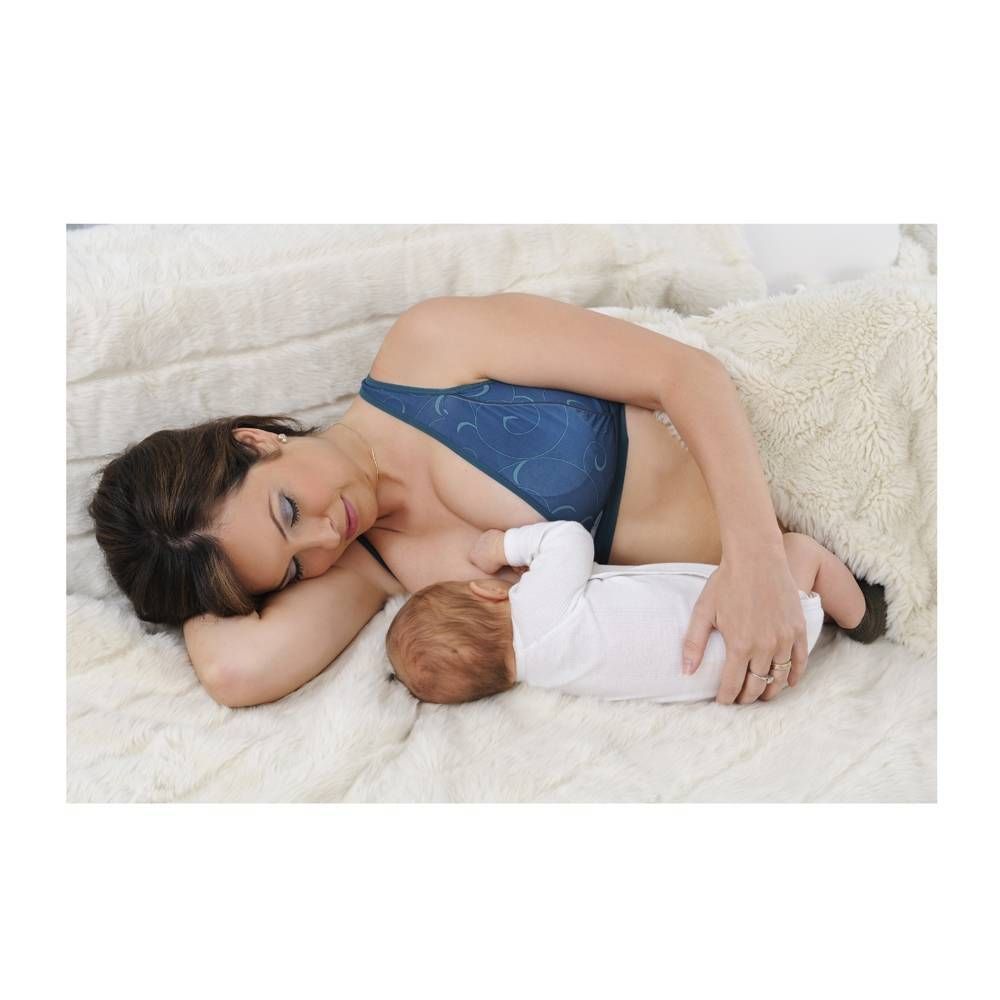 Donna allatta bambino, indossa reggiseno allattamento blu. Sdraiata su pelliccia bianca. Bambino in tutina bianca.
