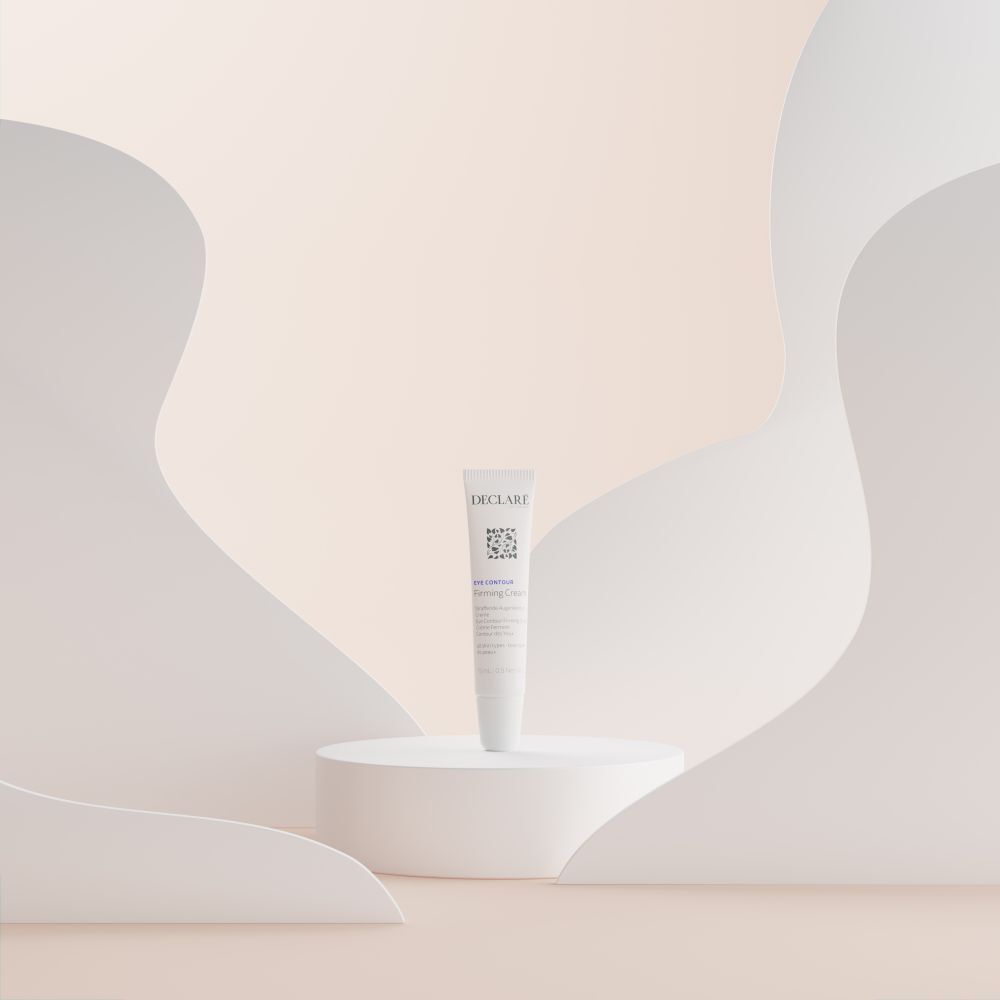 Tubo bianco DECLARÉ Eye Contour Firming Cream su piedistallo bianco. Sfondo con forme bianche.