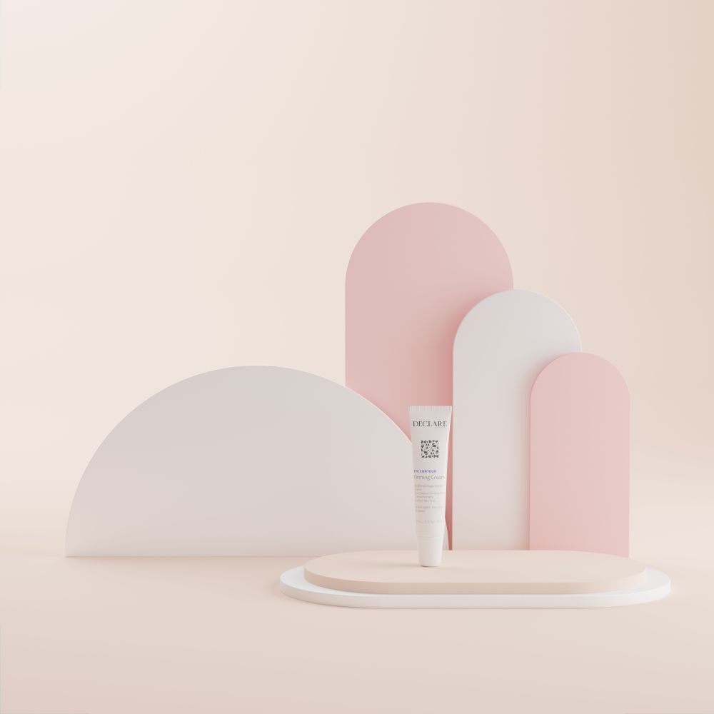 Tubo bianco DECLARÉ Eye Contour Firming Cream su piedistallo. Sfondo con forme rosa e bianche.