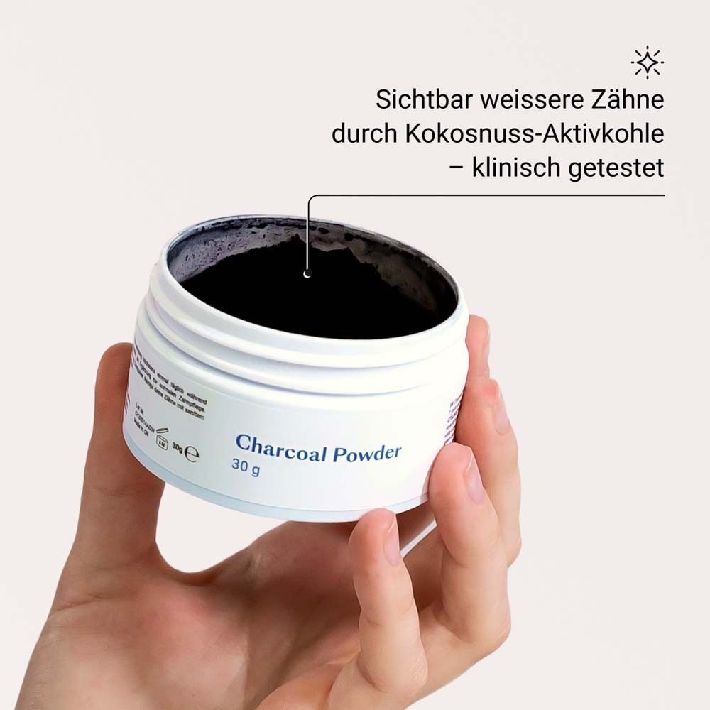 Mano che tiene un barattolo di "Charcoal Powder". Polvere nera visibile. Testo: Denti più bianchi grazie al carbone attivo di cocco.