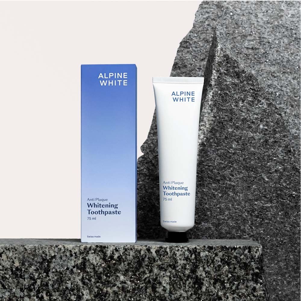 Tubo e scatola di dentifricio. Scatola blu e tubo bianco su pietra. Testo: ALPINE WHITE, Anti-Plaque, Whitening Toothpaste.