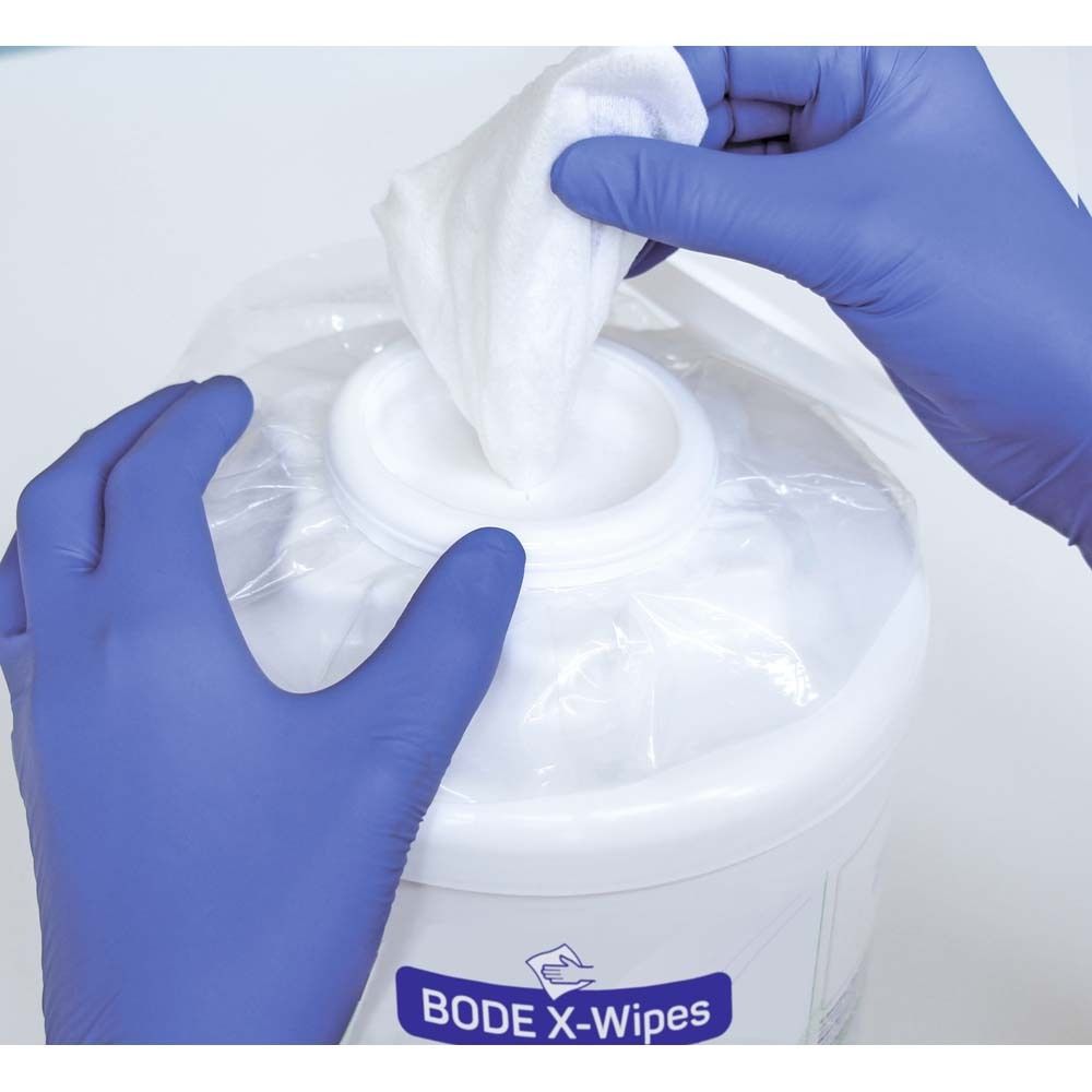 Salviette bianche vengono prelevate da un contenitore bianco. Mani con guanti blu. Sul contenitore è scritto BODE X-Wipes.