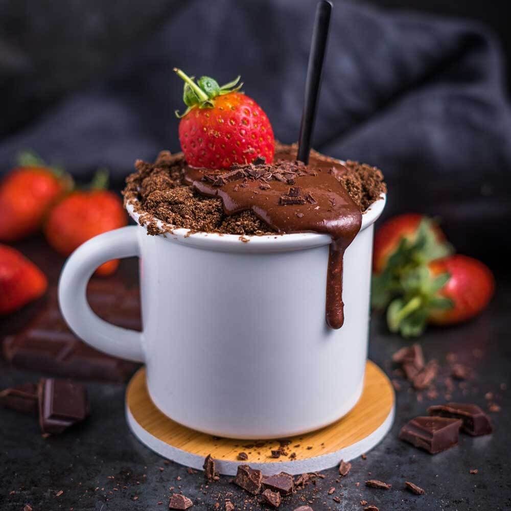 Tazza con torta al cioccolato, fragola, scaglie di cioccolato. Su sottobicchiere di legno.