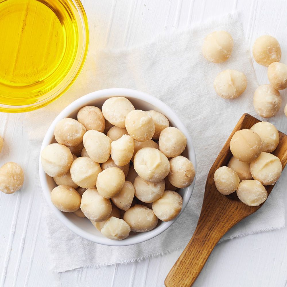 Ciotola bianca con noci di macadamia, cucchiaio di legno con noci, accanto olio in una ciotola di vetro.