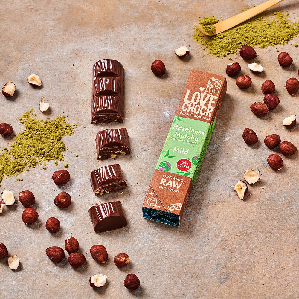 Barretta e pezzi di cioccolato, nocciole e polvere di matcha su sfondo. Confezione: LOVECHOCK, Haselnuss Matcha Mild, Organic Raw Chocolate.