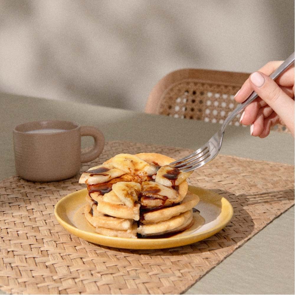 Pancake con fette di banana su un piatto. Una mano tiene una forchetta. Una tazza sullo sfondo.