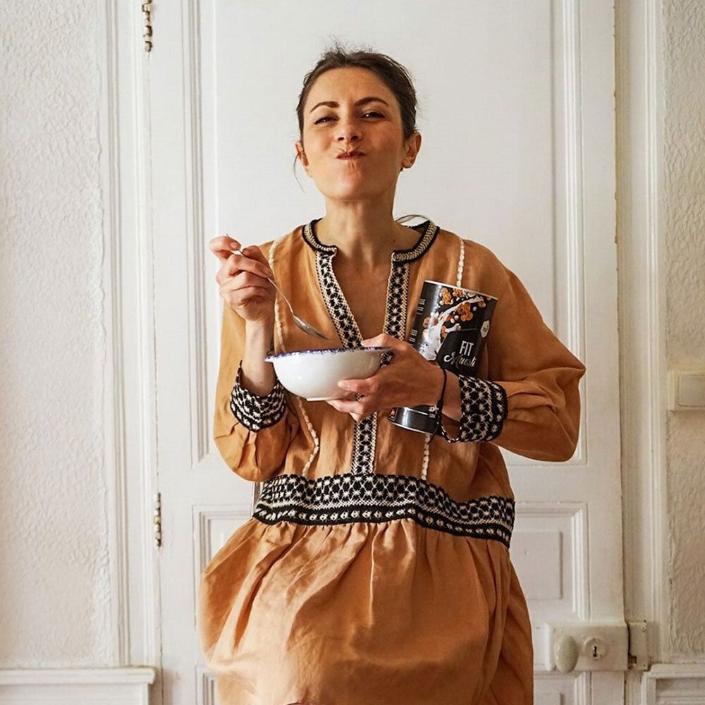Donna con abito beige tiene barattolo nu3 Fit Müsli, Coconut Crunch e mangia da una ciotola. Porta sullo sfondo.