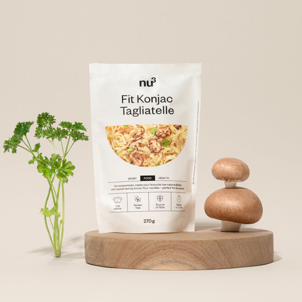 Una confezione di nu3 Fit Konjak-Tagliatelle su un piedistallo di legno, accanto a prezzemolo fresco e funghi.