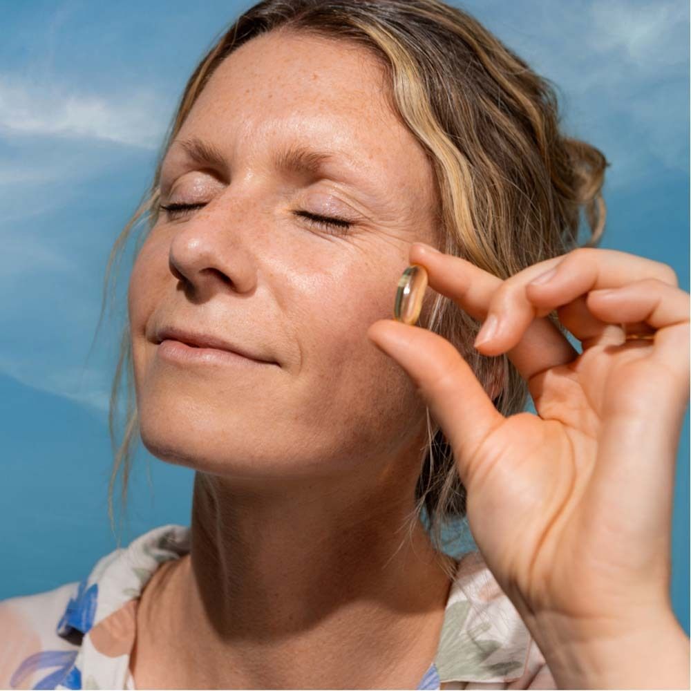 Donna tiene in mano una capsula omega-3 dorata. Cielo blu sullo sfondo.