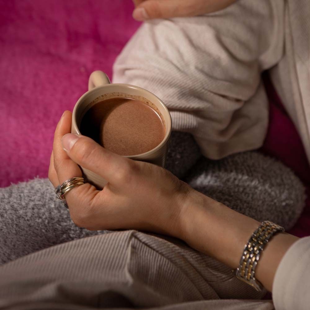 Mani che tengono una tazza di cacao. La persona indossa anelli e un braccialetto. Sfondo: coperta rosa.