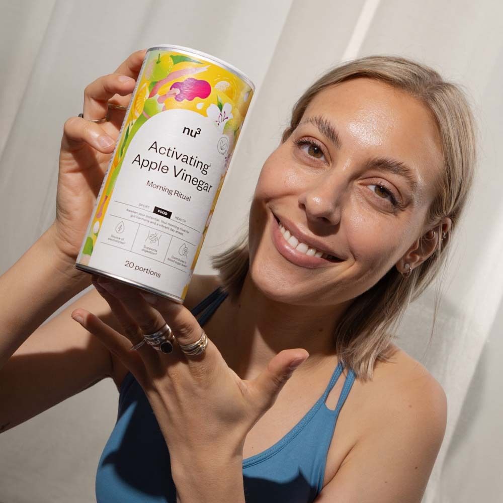 Una donna tiene in mano un barattolo "Activatin Apple Vinegar" verso la fotocamera. Scritta "Morning Ritual". Logo vegano.
