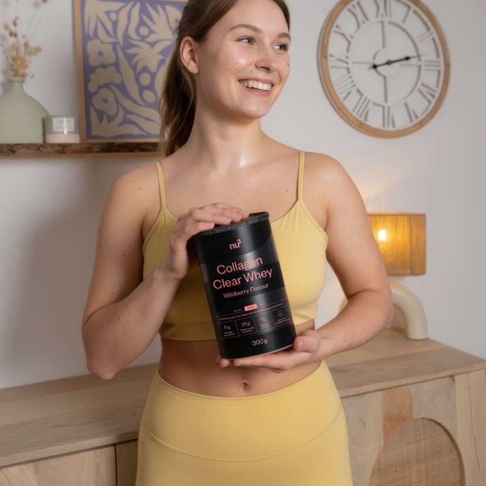 Donna tiene in mano un barattolo nero nu3 Collagen Clear Whey. Abbigliamento sportivo giallo. Sfondo: stanza con orologio e mobili.