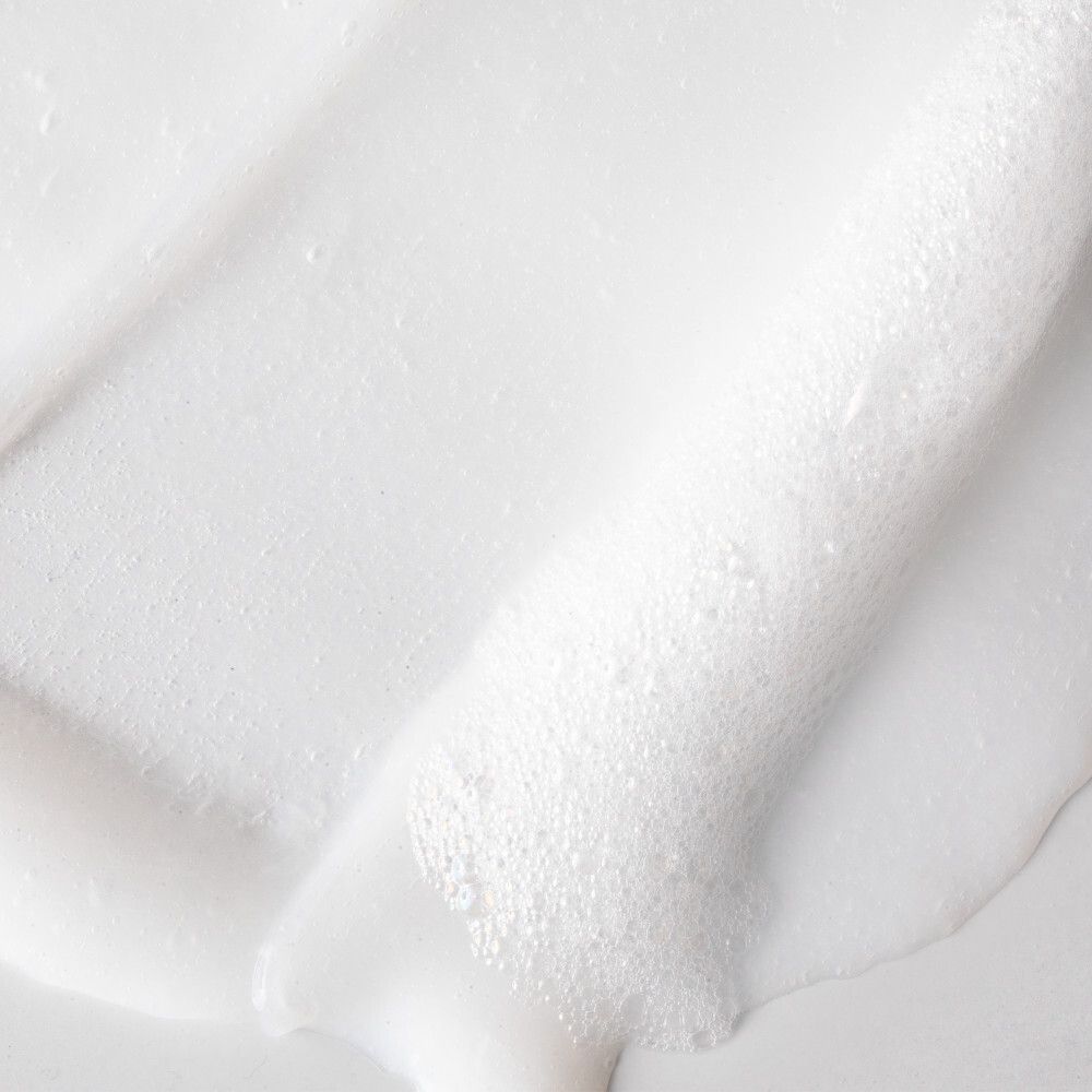 Una mano applica una crema bianca sul viso di una persona. "Formula non schiumogena" è scritto su sfondo blu.