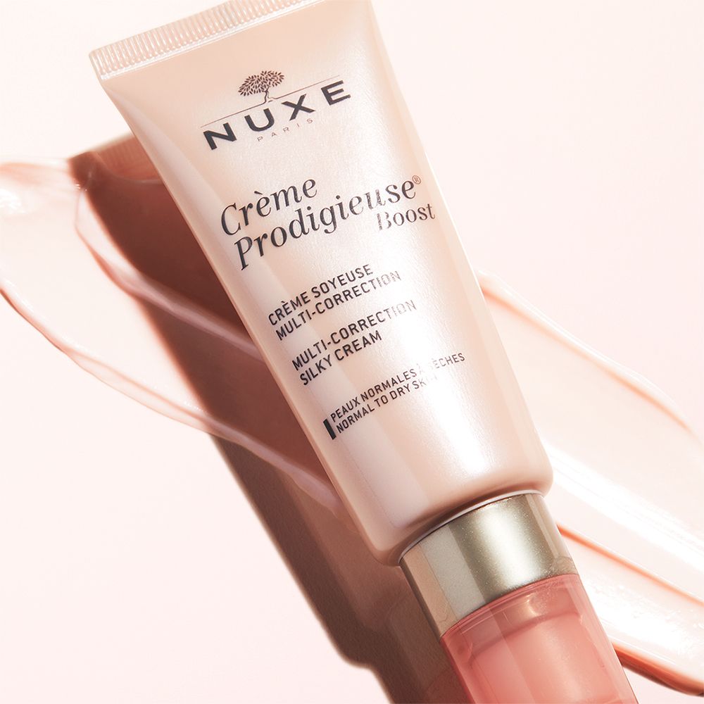 Tubo rosa con crema. Scritta: Nuxe, Crème Prodigieuse Boost. Traccia di crema sullo sfondo.
