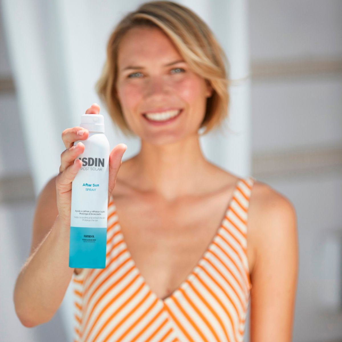 Donna che tiene in mano un flacone spray bianco. ISDIN After Sun Spray. La donna sorride.
