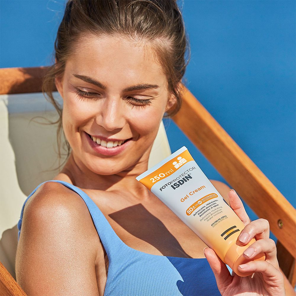Donna tiene in mano tubo ISDIN Fotoprotector Gel Cream LSF 50+. Sorride. Sfondo: spiaggia e mare.