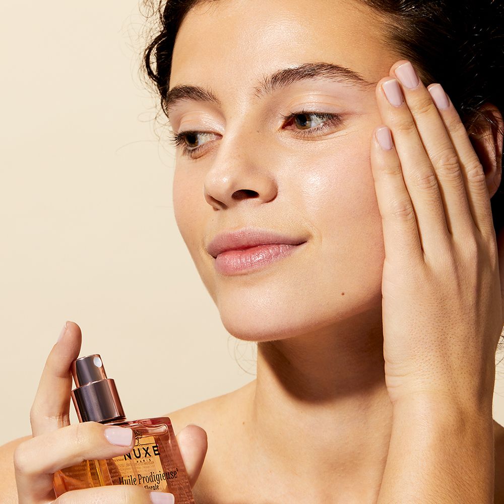 Donna tiene flacone. Scritta: Huile Prodigieuse. Donna tocca il suo viso.