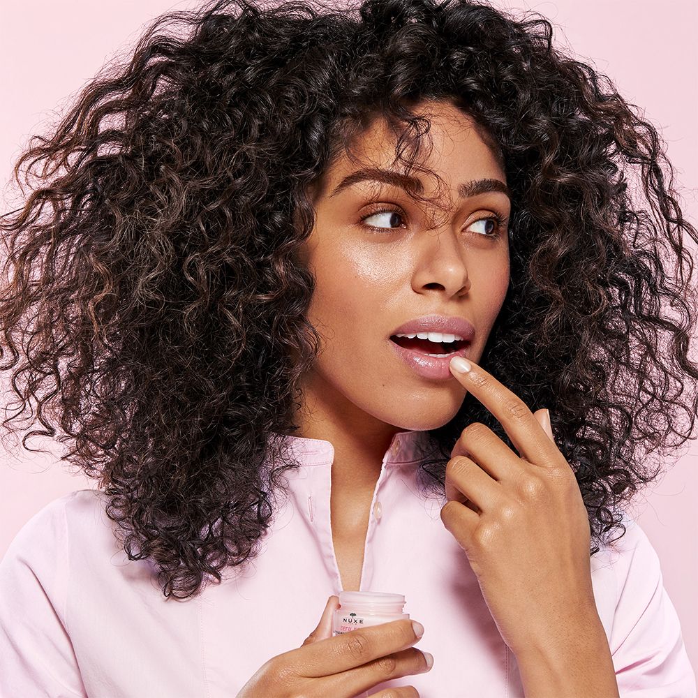 Donna con capelli ricci. Si tocca le labbra con un dito. Piccolo vasetto di balsamo labbra.