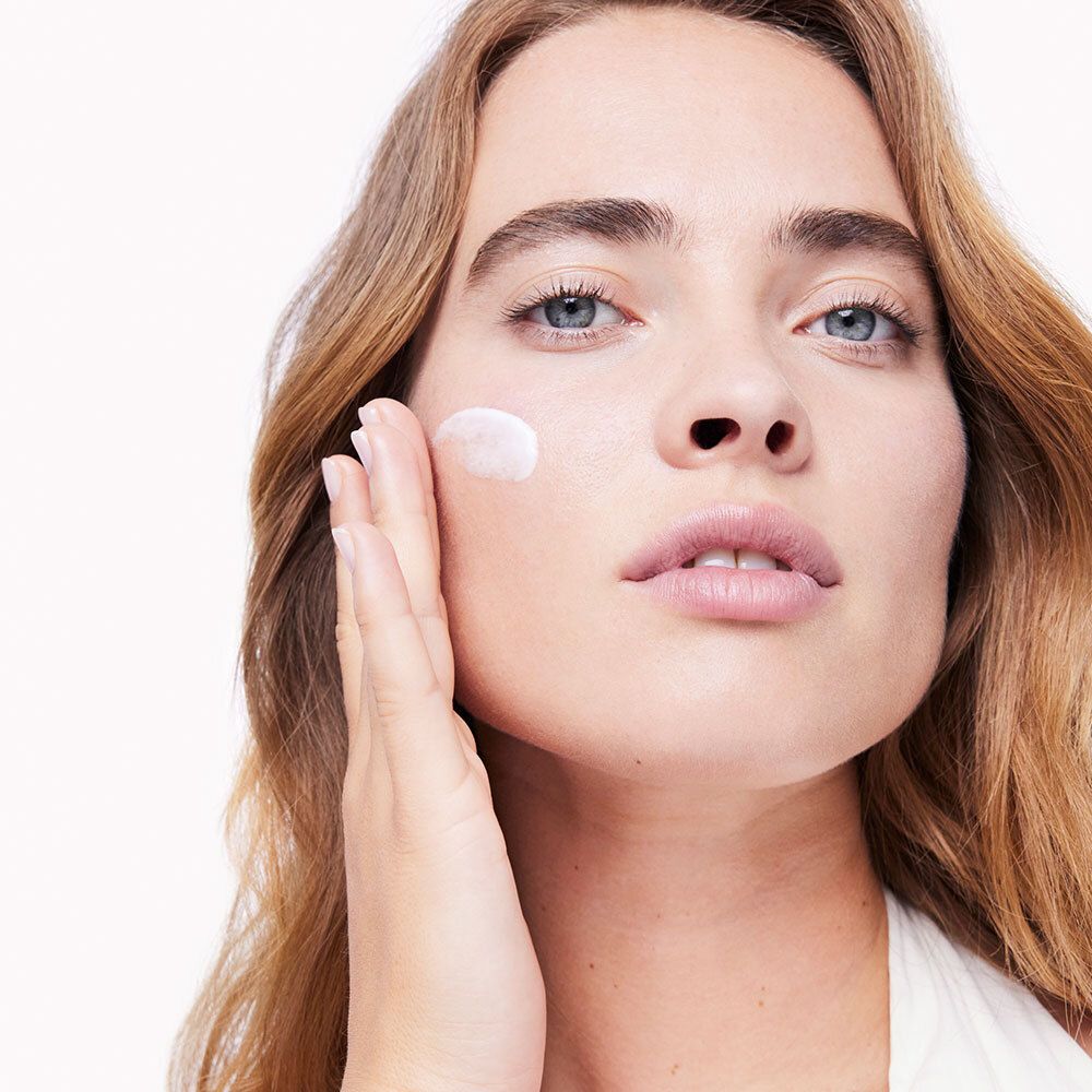 Donna che applica la crema sulla guancia. Pelle chiara, capelli biondi, top bianco.