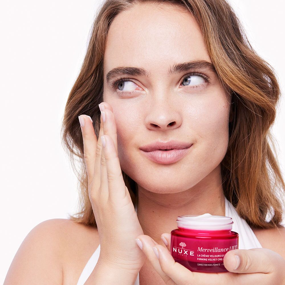 Donna che tiene un vasetto di crema rosso. Applica la crema sul viso. Scritta: Merveillance Lift, NUXE.