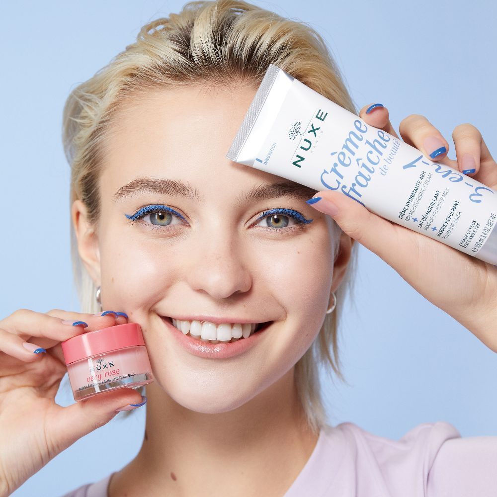 Donna che tiene tubetti di crema e balsamo labbra. Scritta: Crème fraîche de beauté, NUXE. Eyeliner e unghie blu.
