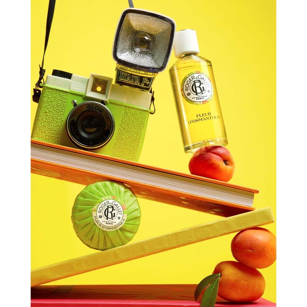 Scena gialla con fotocamera, libri, pesche e sapone. Flacone: Fleur d'Osmanthus.