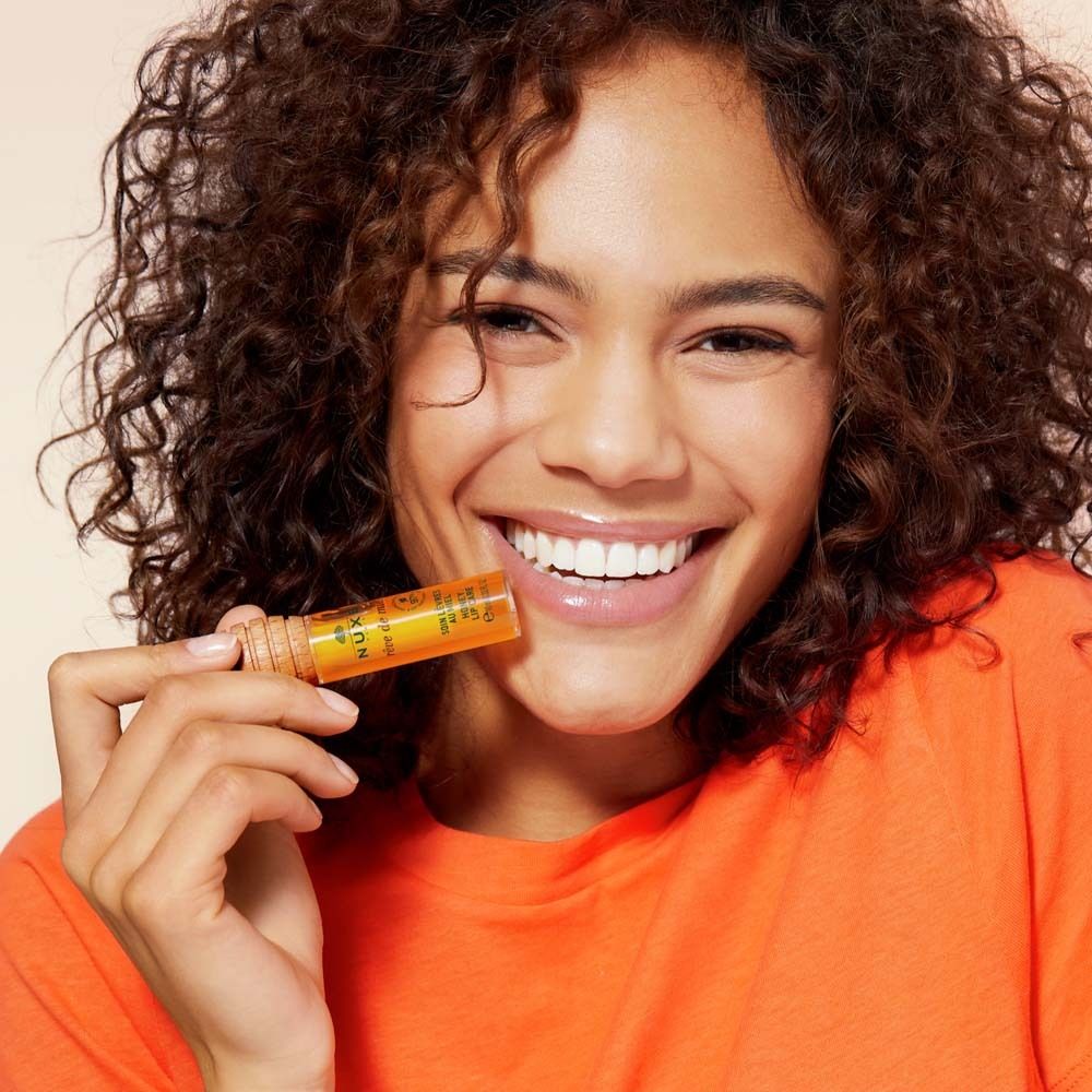 Donna con olio per labbra. Maglietta arancione. Prodotto: NUXE Rêve de Miel®. Tappo in legno visibile.