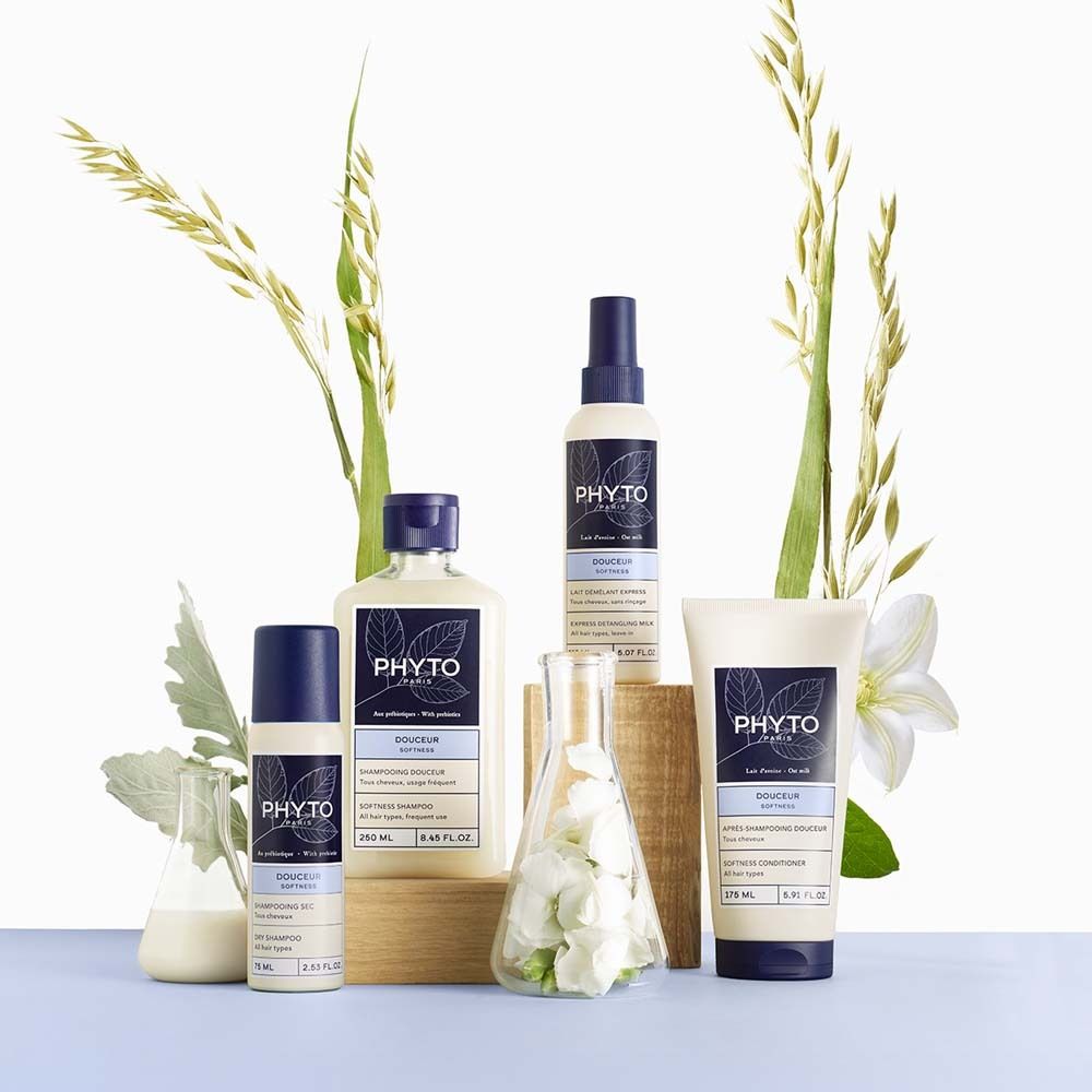 Cinque prodotti per la cura dei capelli Phyto in flaconi e tubetti bianchi con tappi ed etichette blu. Spighe di cereali e fiori sono disposti attorno ai prodotti.