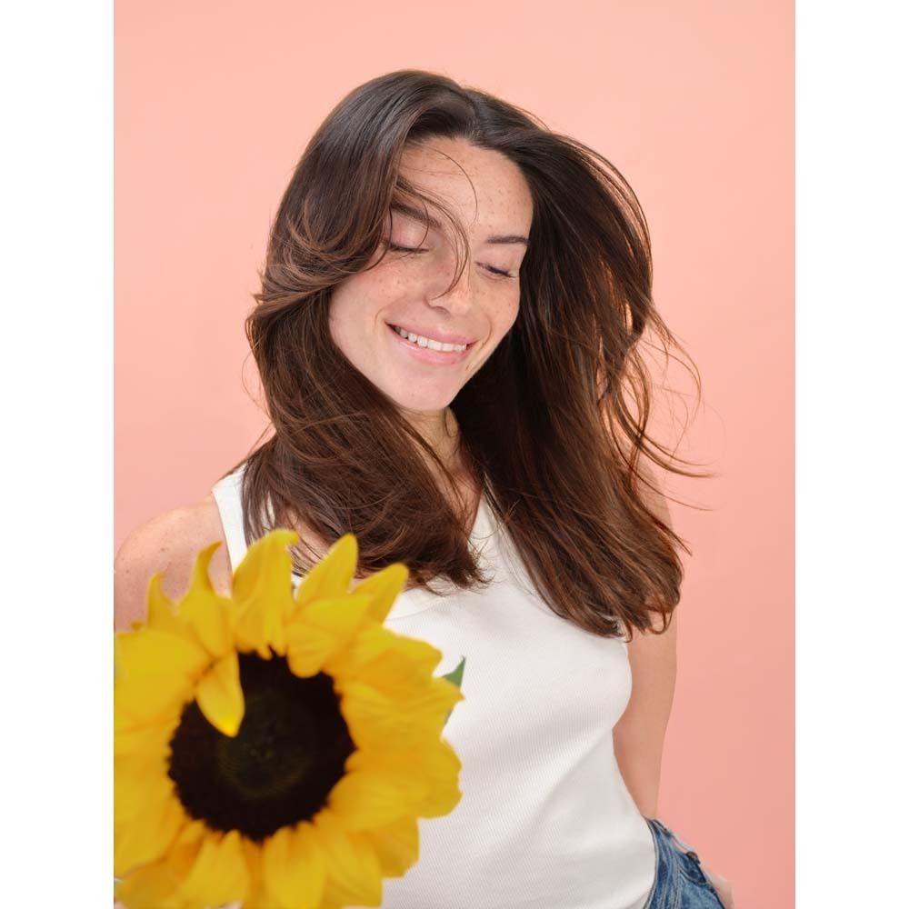 Donna con capelli castani tiene un girasole, indossa un top bianco e jeans.