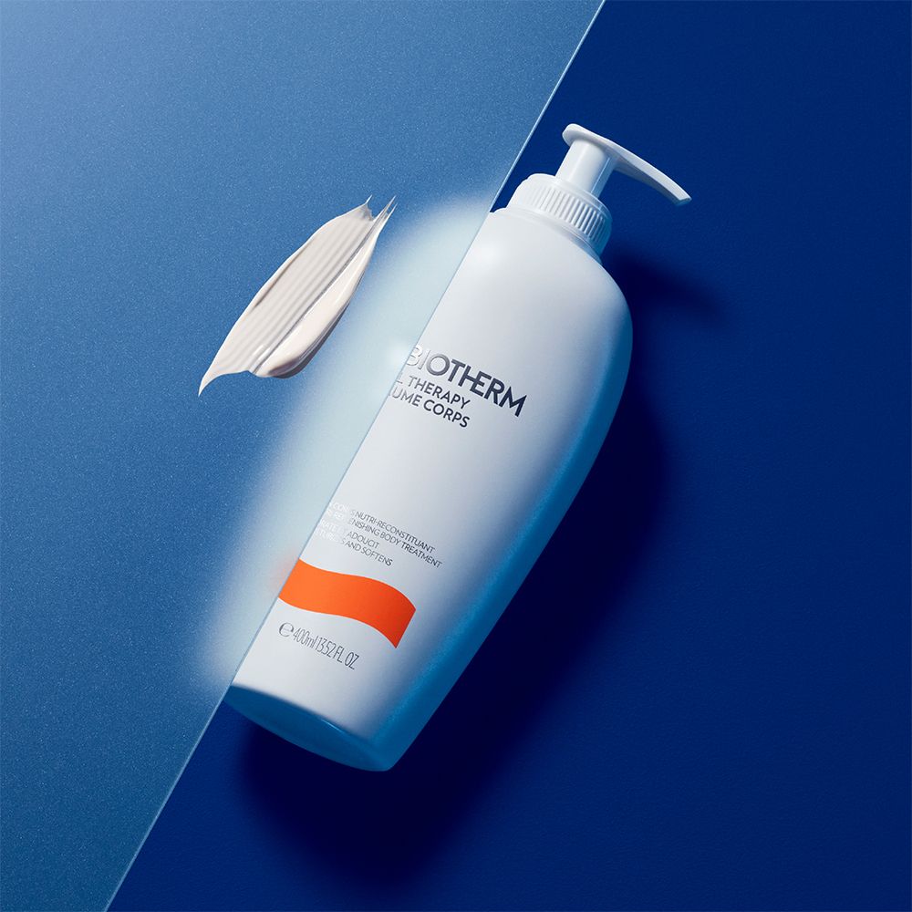 Flacone bianco con dosatore. Testo: BIOTHERM OIL THERAPY BAUME CORPS. Macchia di crema accanto. Sfondo blu.