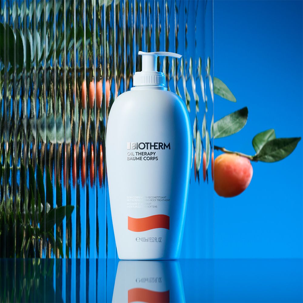 Flacone bianco con dosatore. Testo: BIOTHERM OIL THERAPY BAUME CORPS. Striscia arancione. Sfondo: vetro e frutta.