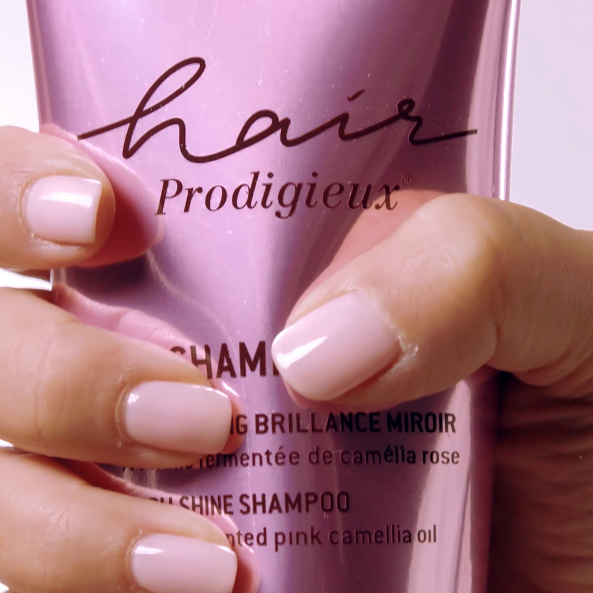 Primo piano di una mano con unghie rosa che tiene un tubetto di shampoo. Testo: Hair Prodigieux.