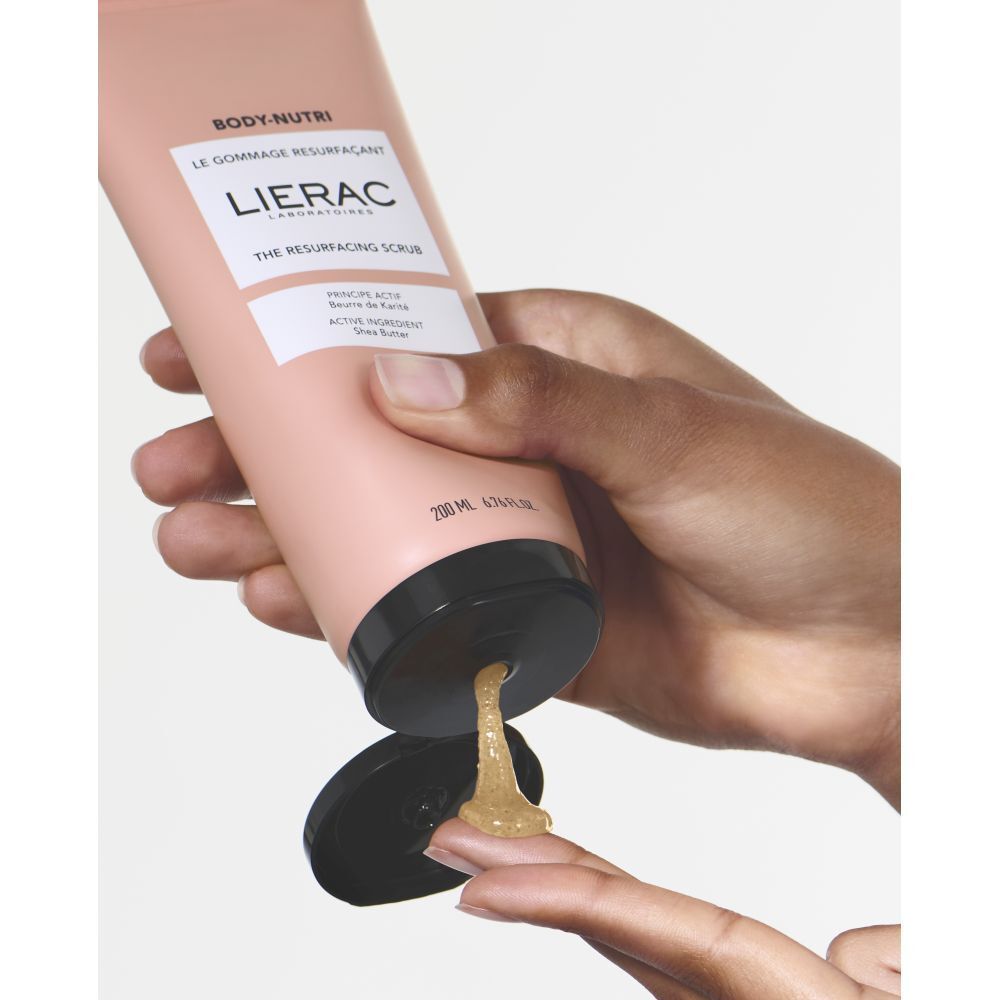 Persona che applica lo scrub sul dito. Tubo rosa con tappo nero. Scritta: LIERAC, BODY-NUTRI, THE RESURFACING SCRUB.