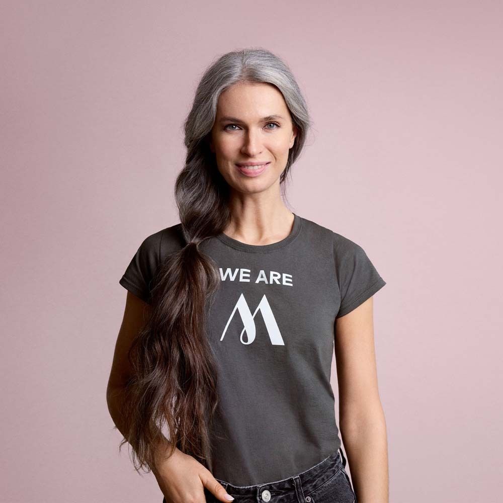 Donna con capelli grigi e maglietta grigia con logo bianco. Scritta: WE ARE M.