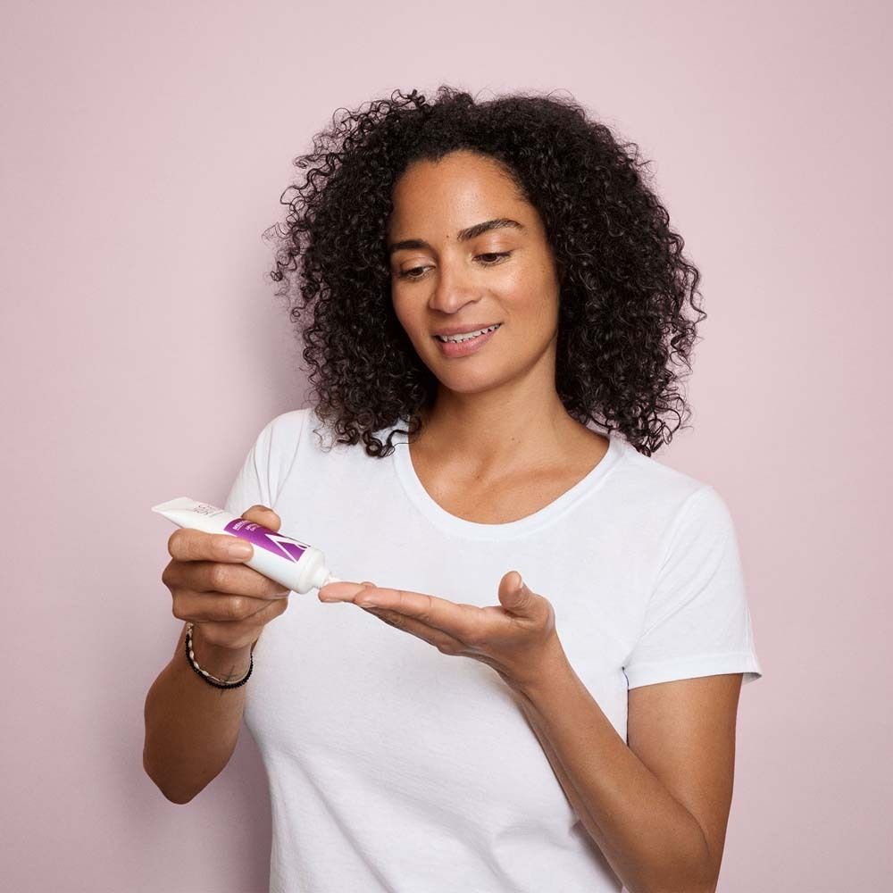 Donna con capelli ricci tiene un tubetto. Spreme il prodotto sulla mano. Sfondo rosa.