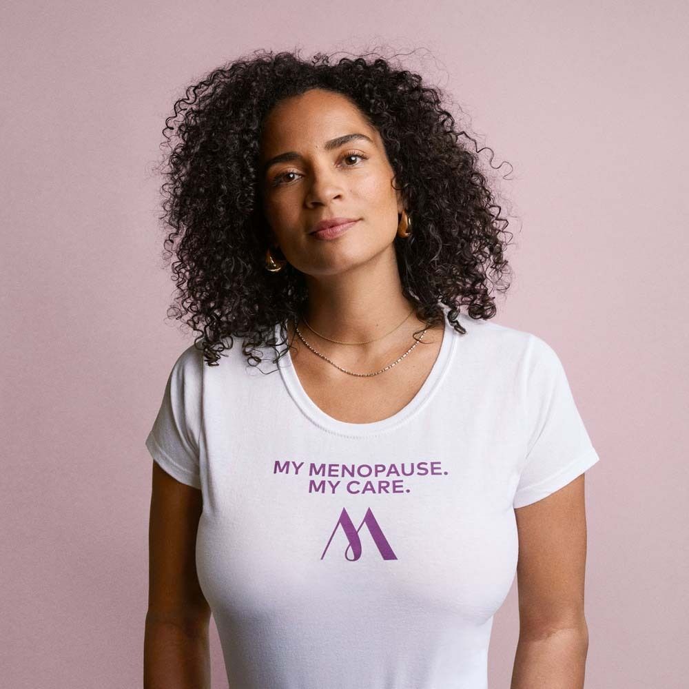Donna con maglietta bianca. Stampa: My Menopause. My Care. Logo: M viola. Sfondo rosa.