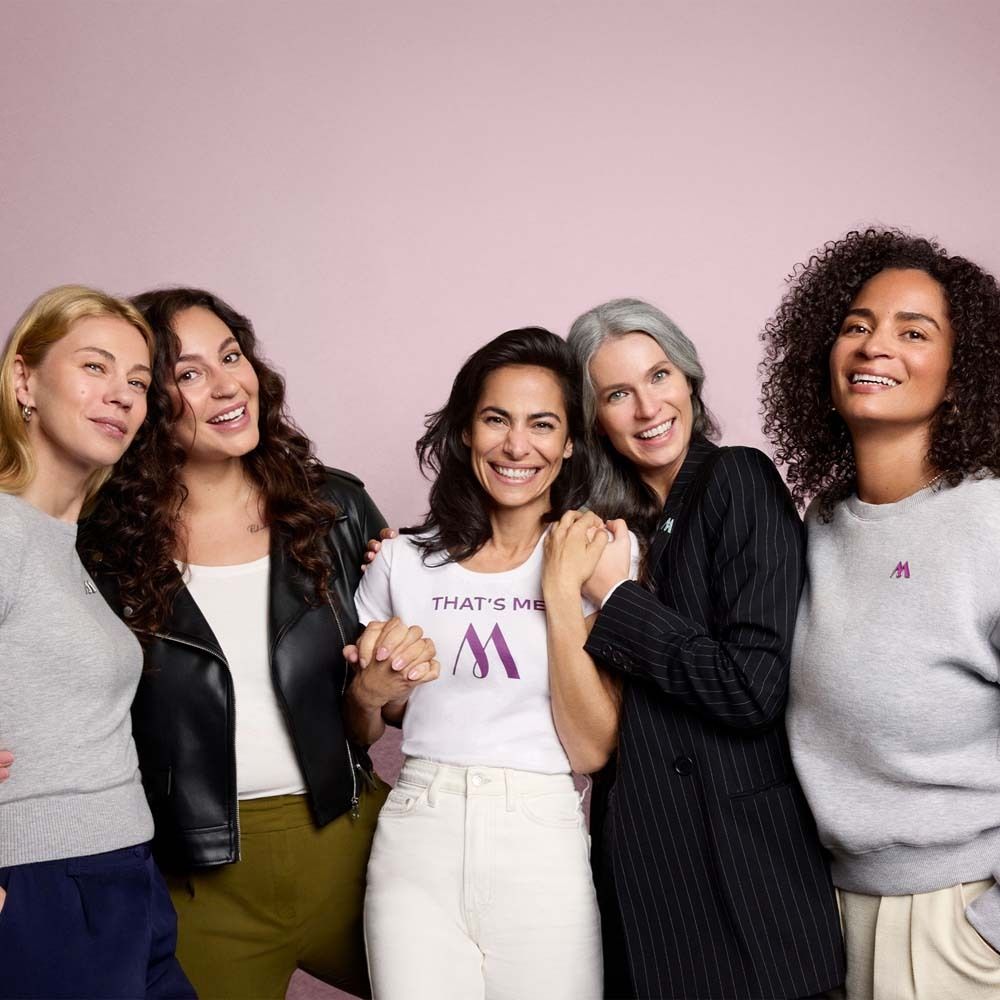 Cinque donne in piedi fianco a fianco. Sorridono. Una indossa una maglietta con logo.